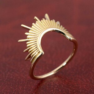 May include: A gold ring with a sunburst design. The ring has a curved band with a half-circle sunburst design on top, with multiple rays extending outwards. The ring is set against a red background.
