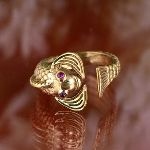 May include: A gold-colored ring shaped like a fish, featuring a detailed design with scales and fins. The fish has two small, round, red gemstones for eyes. The ring is open at the back, allowing for size adjustment.