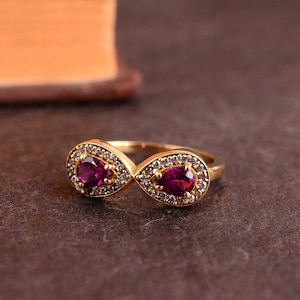 Può includere: Un anello color oro con un design a simbolo dell'infinito. Ogni anello del simbolo dell'infinito presenta una gemma rosso scuro a forma di goccia, circondata da piccole pietre trasparenti. L'anello è posto su uno sfondo marrone scuro.