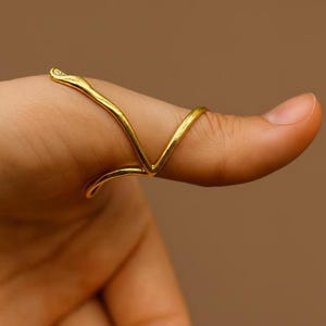 Peut inclure: Une bague dorée au design abstrait unique. La bague épouse le doigt, avec une forme en V et un petit élément décoratif en haut. La bague est présentée sur un doigt, sur fond marron.