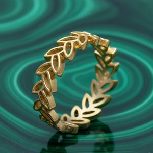May include: A gold ring with a repeating leaf pattern. The ring's leaves are finely detailed, giving it a delicate and elegant appearance. The ring is set against a green background.