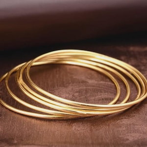 Puede incluir: Un conjunto de seis pulseras doradas, cada una con un diseño simple y redondeado. Las pulseras están apiladas, creando un efecto de capas. Las joyas se exhiben sobre una superficie marrón.