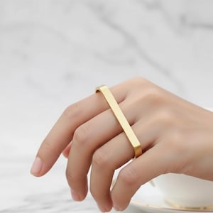 Puede incluir: Un anillo dorado con un diseño de barra rectangular. El anillo se lleva en un dedo, mostrando su estilo simple pero elegante. La superficie lisa del anillo refleja la luz, resaltando su acabado metálico.