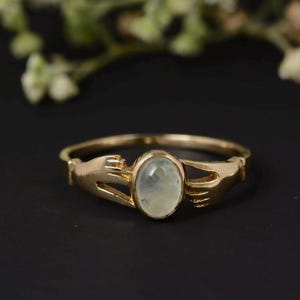 May include: A gold ring with a light green oval gemstone. The ring's design features two hands holding the gemstone. The ring is set against a black background, with blurred greenery in the background.