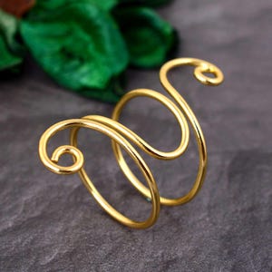 May include: A gold-colored ring with a unique, open design. The ring features a swirling, wire-like structure with two main loops and decorative spirals at the ends. The jewelry is set against a dark gray background with a hint of green.