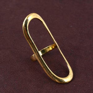 Puede incluir: Un anillo dorado con un diseño ovalado largo y abierto. El anillo tiene una banda que rodea el dedo y una barra de conexión dentro del óvalo. El anillo está sobre un fondo granate oscuro.