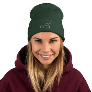 Może przedstawiać: Ciemnozielona czapka typu beanie z białą, haftowaną strukturą chemiczną z przodu. Czapka wykonana jest z miękkiego materiału i ma wywinięty mankiet. Osoba na zdjęciu ma na sobie bordową bluzę z kapturem.
