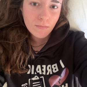 Könnte beinhalten: Eine Person trägt einen schwarzen Hoodie mit dem weißen Text "I Break Hearts Not Again" und einer roten, zerbrochenen Herzgrafik. Die Person hat braune Haare und liegt.
