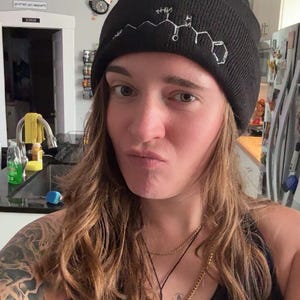 Puede incluir: Gorro negro con un diseño de estructura química blanca. El gorro está hecho de un material suave y tejido. La persona de la imagen lleva una camiseta sin mangas negra y un collar con un colgante de espada.