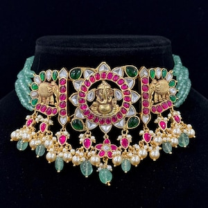 May include: An ornate gold-toned necklace featuring a central pendant with a deity figure, surrounded by red and clear gemstones. The necklace includes green and white beads, with dangling pearl and gemstone accents. The design incorporates elephant motifs.