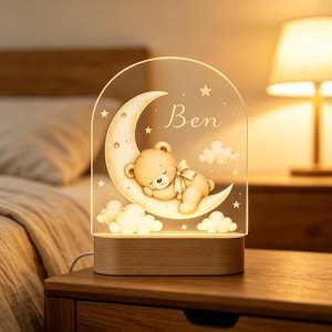Könnte beinhalten: Ein bogenförmiges Acryl-Nachtlicht mit Holzsockel, das eine Illustration eines hellbraunen Teddybären zeigt, der auf einer Mondsichel zwischen Wolken und Sternen schläft, wobei der Name Ben in Schreibschrift über dem Bären gedruckt ist.