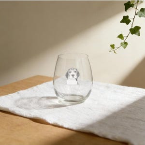 Peut inclure: Un verre à vin sans pied transparent avec un portrait de beagle en noir et blanc. Le verre est posé sur un tissu blanc, avec une table en bois et une plante verte en arrière-plan. Le verre est de forme arrondie.
