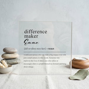 Könnte beinhalten: Klarer Acrylblock mit den schwarzen Aufdrucken "difference maker Emma". Die Definition von "difference maker" ist ebenfalls auf dem Block aufgedruckt. Ein Stapel Steine und eine kleine Schale befinden sich im Hintergrund.