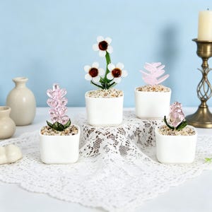 Könnte beinhalten: Vier dekorative Glasblumenarrangements in weißen quadratischen Töpfen. Die Blumen sind in verschiedenen Farben, darunter Rosa und Weiß, und sitzen auf einem Bett aus kleinen Steinen. Die Töpfe sind auf einer weißen Spitzentischdecke angeordnet.