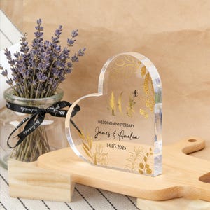 Könnte beinhalten: Ein herzförmiges Jubiläumandenken aus klarem Acryl mit goldenen floralen Akzenten. Der Text lautet "40. Hochzeitstag" und die Namen "James & Amelia" mit dem Datum 14.03.2025. Ein Glas Lavendel befindet sich im Hintergrund.