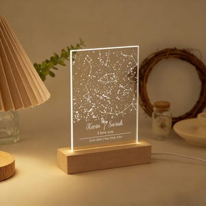 Könnte beinhalten: Personalisiertes Nachtlicht mit Sternbildkarte. Die transparente Acrylplatte zeigt eine Sternenkarte mit den Namen "Kevin & Sarah" und dem Text "I love you". Es steht auf einem Holzsockel und strahlt ein warmes Licht aus.