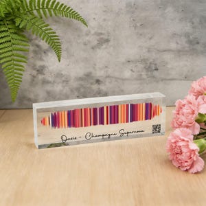 Könnte beinhalten: Ein klarer Acrylblock mit einem farbenfrohen, vertikalen Streifendesign in Orange, Lila und Rot. Der Text "Oasis - Champagne Supernova" ist auf der Vorderseite aufgedruckt. Ein QR-Code ist auf der rechten Seite sichtbar.