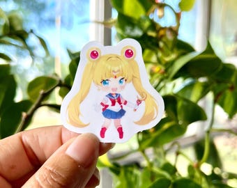 May include: A die-cut vinyl sticker featuring a chibi-style anime character with long blonde hair in twin tails, wearing a blue and white sailor-style uniform with a red bow, red boots, and holding a small pink wand with a star-shaped top.