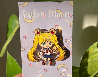 May include: A metal enamel pin featuring a chibi-style Sailor Moon character with blonde hair, blue eyes, and a red, white, and blue uniform. The pin is attached to a light purple backing card decorated with yellow crescent moons, stars, and white rabbits.