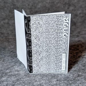 May include: A small, open book with white pages and a black and white cover. The cover has a textured pattern and the year "2026" is printed on the top right corner. The spine has vertical lines and text.