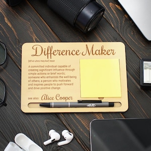 May include: A wooden desk organiser with the text "Difference Maker" and a definition. Includes a yellow sticky note, a pen, and a smartphone. Other items include a camera, glasses, and wireless earbuds.