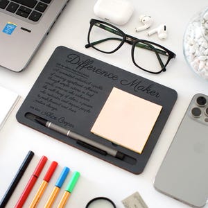 Puede incluir: Un organizador de escritorio gris oscuro con el texto "Difference Maker" y una definición. Incluye un bolígrafo, una nota adhesiva y una ranura para un teléfono. Otros artículos incluyen una computadora portátil, gafas y un teléfono inteligente.