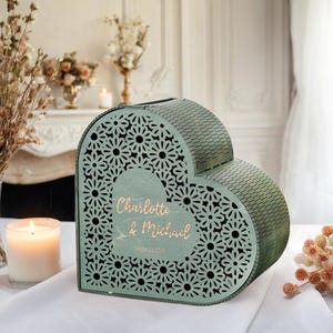 May include: A sage green heart-shaped card box with a floral cut-out design. The box has a hinged lid and features the names "Charlotte & Michael" in gold script. A candle and floral arrangements are in the background.