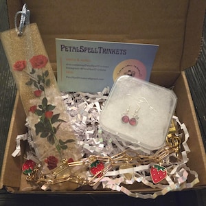 May include: A gift box with a gold glitter bookmark featuring red roses, pink gemstone earrings, and a gold chain bracelet with strawberry charms. The box also contains a business card with the text "Petal Spell Trinkets".