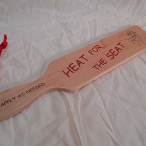 May include: A wooden paddle with a red rope handle. The paddle is engraved with the text "HEAT FOR THE SEAT" and "APPLY AS NEEDED".