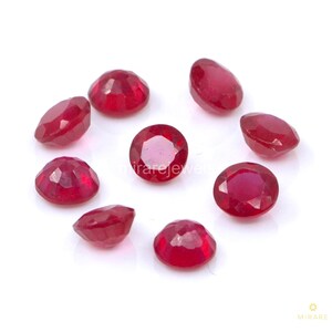 Könnte beinhalten: Eine Ansammlung facettierter, tiefroter Edelsteine, möglicherweise Rubine, auf einer weißen Oberfläche angeordnet. Die Edelsteine haben unterschiedliche Formen, einige rund, andere oval. Die Facetten fangen das Licht ein und erzeugen einen funkelnden Effekt. Das Bild ist gut beleuchtet und hebt die Farbe und Klarheit der Steine hervor.