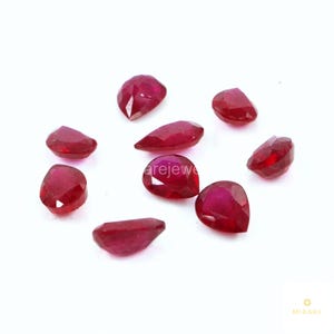 May include: A collection of nine loose, faceted rubies in various shapes including pear, oval, and marquise cuts. These gemstones exhibit a deep, vibrant red colour with reflective surfaces and clear, polished facets arranged on a plain white background.