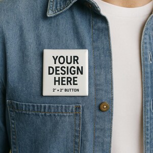 May include: A white, square 2" x 2" button with the text "YOUR DESIGN HERE" in black, pinned to a denim shirt. The button is centered on the shirt, above the pocket.