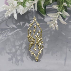 Puede incluir: Un anillo dorado con un diseño intrincado y calado. El anillo presenta una serie de líneas curvas y pequeños acentos esféricos, creando una apariencia delicada y artística. El anillo se muestra sobre una superficie gris con acentos florales blancos en el fondo.