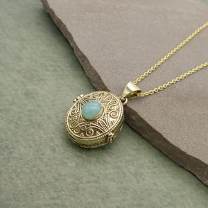 Puede incluir: Un collar con colgante dorado con una piedra preciosa ovalada de color azul claro en el centro. El colgante redondo tiene un diseño ornamental y arremolinado. La cadena del collar es delicada y dorada.