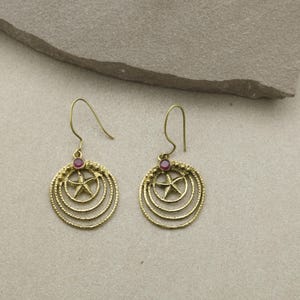 May include: A pair of gold-colored earrings. Each earring features a circular design with a star in the center, surrounded by concentric circles. A small, round, red gemstone is set at the top of each earring.