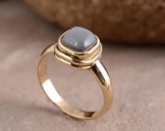 May include: A gold-colored ring with a square, gray gemstone. The ring has a simple band and a decorative setting around the stone. The gemstone is a muted gray color, and the ring appears to be made of metal.