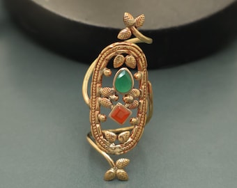 May include: Gold-coloured ring with an oval centrepiece. The ring features a green oval gemstone and a red square gemstone. The design includes leaf and floral accents. The ring has a unique, elongated shape.