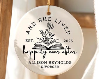May include: A round white ceramic ornament hanging by a gold string. The front features black text reading AND SHE LIVED EST. 2026 happily ever after ALLISON REYNOLDS DIVORCED, with a central graphic of an open book and a flower stem.