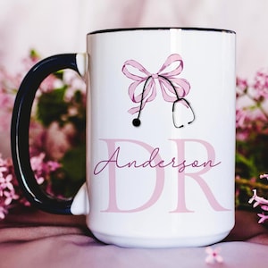 Può includere: Tazza in ceramica bianca con manico e bordo neri. La tazza presenta un design con fiocco e stetoscopio rosa, con il nome "Anderson" e le lettere "DR" in rosa. La tazza è circondata da fiori rosa.