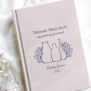 Może przedstawiać: Jasnoróżowy dziennik ceramiczny z napisem "THROWING THINGS HELPS my pottery journal", imieniem "Lacey Gunn" i rokiem "2026". Okładka zawiera niebieską ilustrację ceramiki i liści.