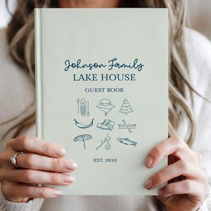 Puede incluir: Un libro de visitas verde claro con el texto "Johnson Family LAKE HOUSE GUEST BOOK" en escritura azul marino. La portada presenta ilustraciones en azul marino con temática de lago y el año "EST. 2010".