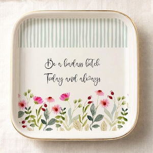 Puede incluir: Un plato cuadrado de cerámica para joyas con borde dorado. La parte superior tiene rayas verticales azul claro, mientras que la inferior muestra un estampado floral en rosa y verde. El texto dice: Be a badass bitch Today and always.