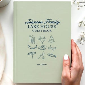 Könnte beinhalten: Ein salbeigrünes Hardcover-Gästebuch mit dunkelblauem Text Johnson Family LAKE HOUSE GUEST BOOK est. 2010. Der Einband zeigt ein Raster aus kleinen Strichzeichnungen, darunter ein Kajak, ein Hut, ein Baum, eine Hängematte und ein Fisch.