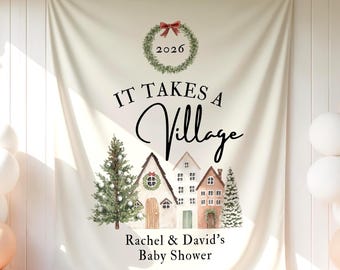 May include: A cream-colored fabric backdrop featuring a watercolor illustration of a winter village with houses and pine trees. The text reads IT TAKES A Village, 2026, and Rachel & David's Baby Shower, topped with a green wreath and red bow.