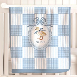 Op de afbeelding: Een babydeken met een blauw en wit geblokt en gestreept patroon. De deken heeft een ovaal ontwerp met een eendenillustratie en de naam "NOAH". De deken is over een wit ledikant gedrapeerd.