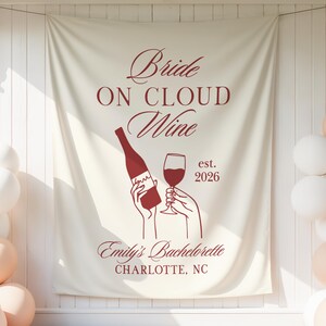 May include: A cream-colored fabric backdrop featuring dark red text that reads Bride ON CLOUD Wine, est. 2026, Emily's Bachelorette, and CHARLOTTE, NC. The design includes a graphic of two hands holding a wine bottle and a glass of red wine.