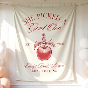 May include: A cream-colored fabric backdrop featuring red text that reads SHE PICKED A Good One, EST. 2026, Emily's Bridal Shower, and CHARLOTTE, NC. A detailed red illustration of an apple with leaves is centered between the text elements.