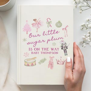 May include: A cream-colored book cover featuring watercolor illustrations of a pink Christmas tree, a ballerina, a candy cane, a nutcracker, a drum, and ballet slippers. The text reads: Our little sugar plum IS ON THE WAY BABY THOMPSON.