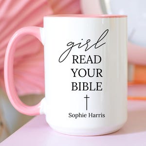 May include: White ceramic mug with a pink handle and interior. The mug features the text "girl READ YOUR BIBLE" in black script, with a cross symbol and the name "Sophie Harris" below. The mug is on a pink surface.
