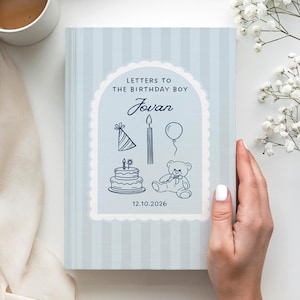 Puede incluir: Un libro de tapa dura con rayas azul claro titulado Letters to the Birthday Boy Jovan, que presenta un gráfico de arco festoneado con ilustraciones de un gorro de fiesta, vela, globo, pastel y oso de peluche, y la fecha 12.10.2026 en tinta azul.